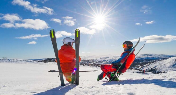 Par som sitter på sina skidor i skidbacken, soligt och snörikt fjällandskap i bakgrunden