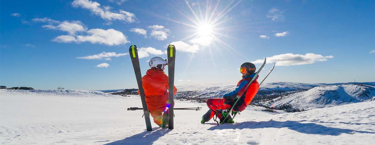 Par som sitter på sina skidor i skidbacken, soligt och snörikt fjällandskap i bakgrunden