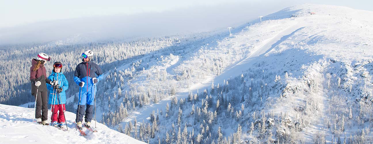 Familj på toppen av skidbacken hovdetoppen iklädda skidkläder med pjäxor, stavar och skidor.